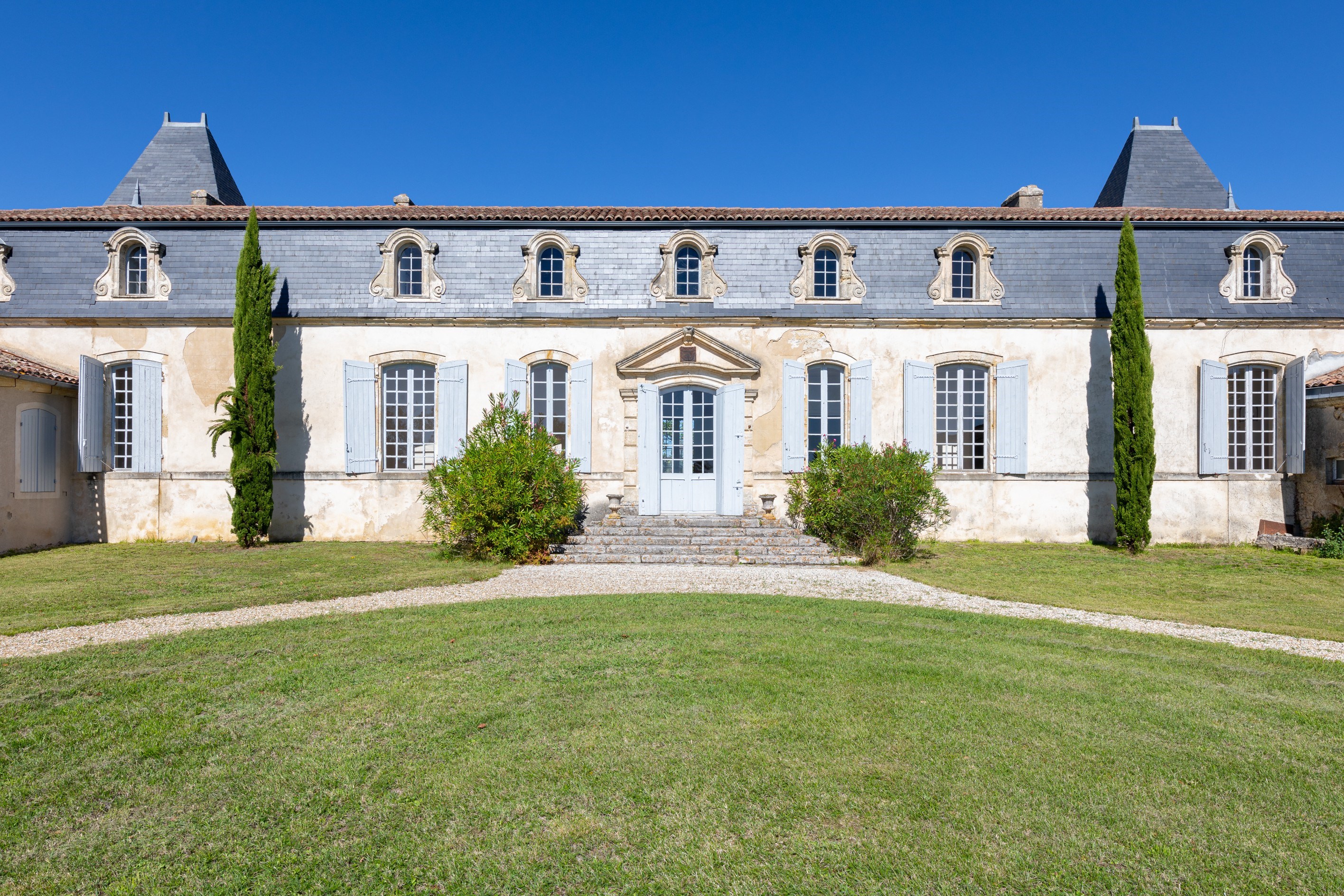 Pale stone manor with blue shutters, slate roof, and cypress trees beside the entrance.