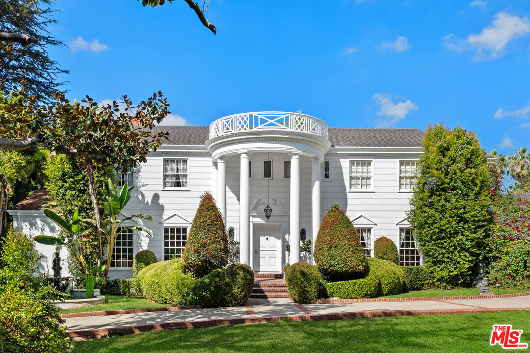 White two-story house with a columned portico, rounded balcony, and formal hedges.