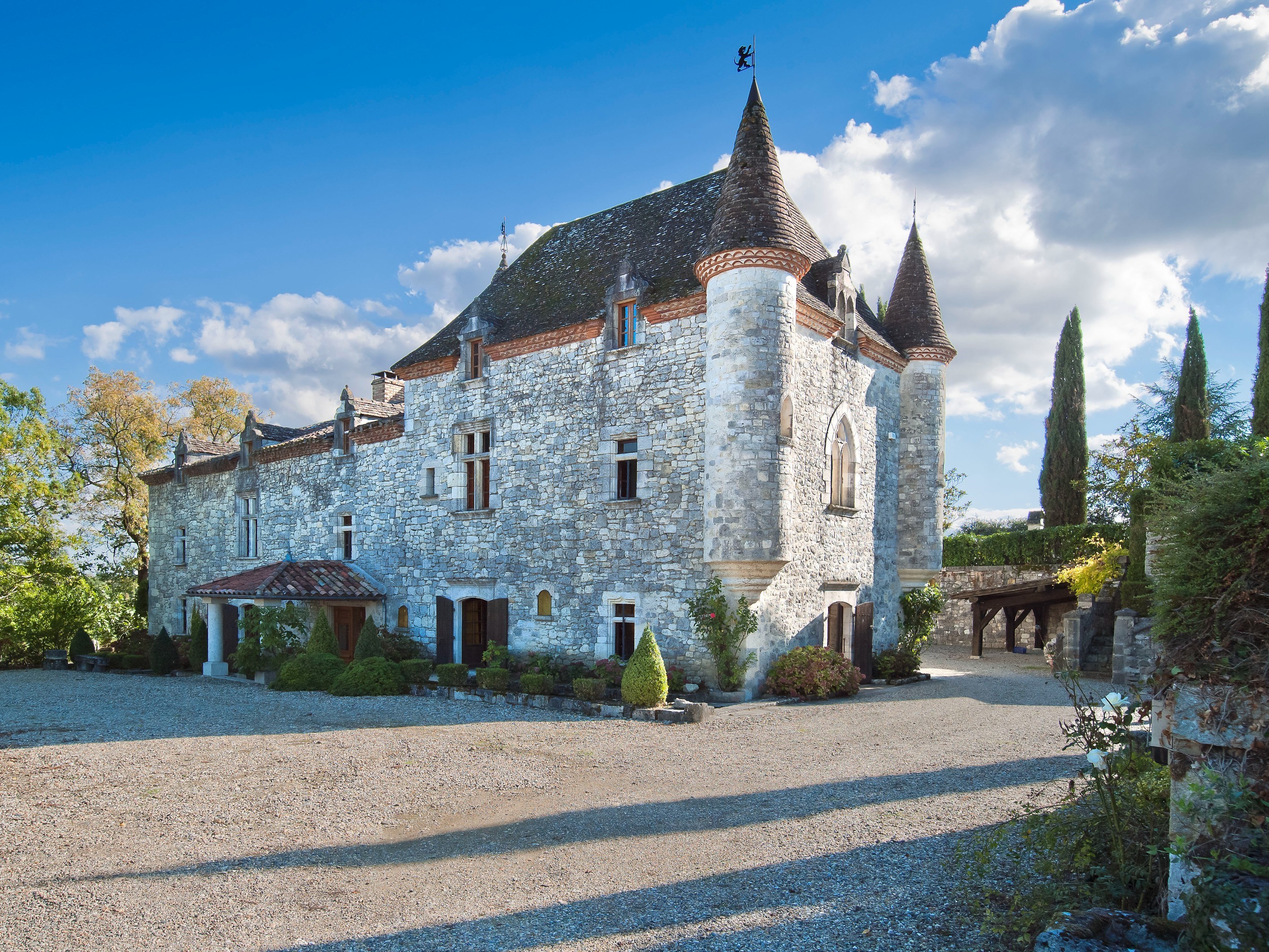 Stone building with conical towers and a steep roof beside a gravel courtyard.
