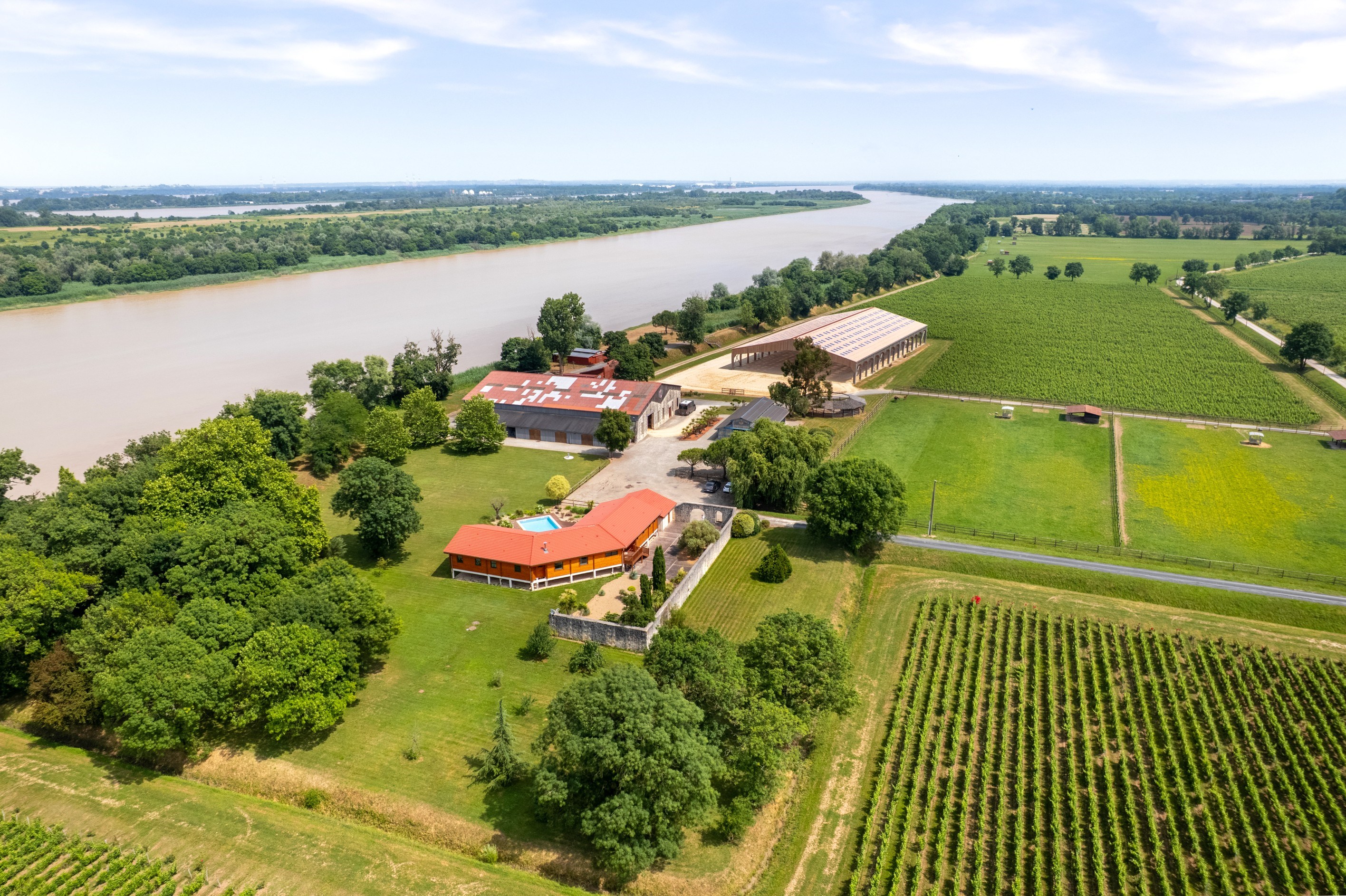 Rural estate beside a river, with a pool, vineyard rows, and red-roofed buildings.