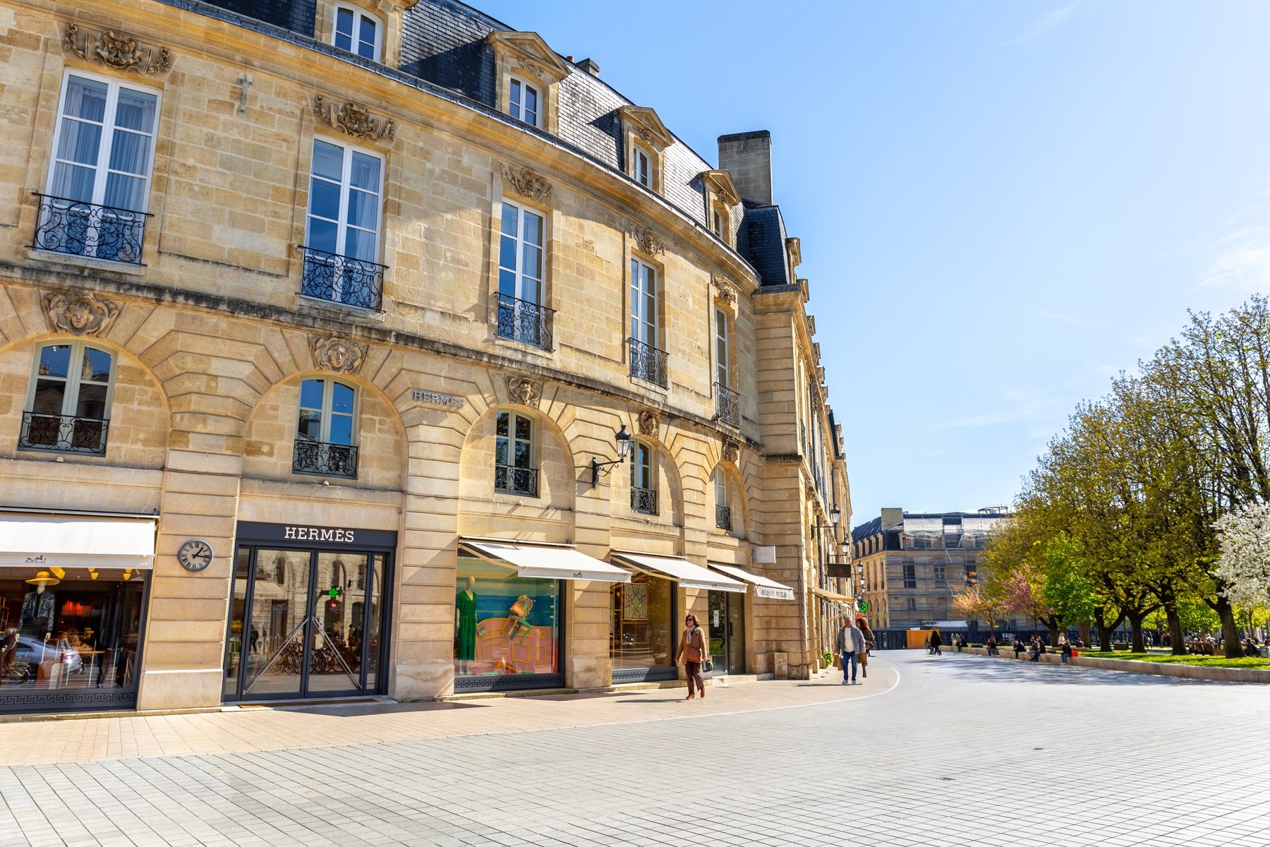 Pedestrian street beside a curved stone building with Hermès storefronts and spring trees.