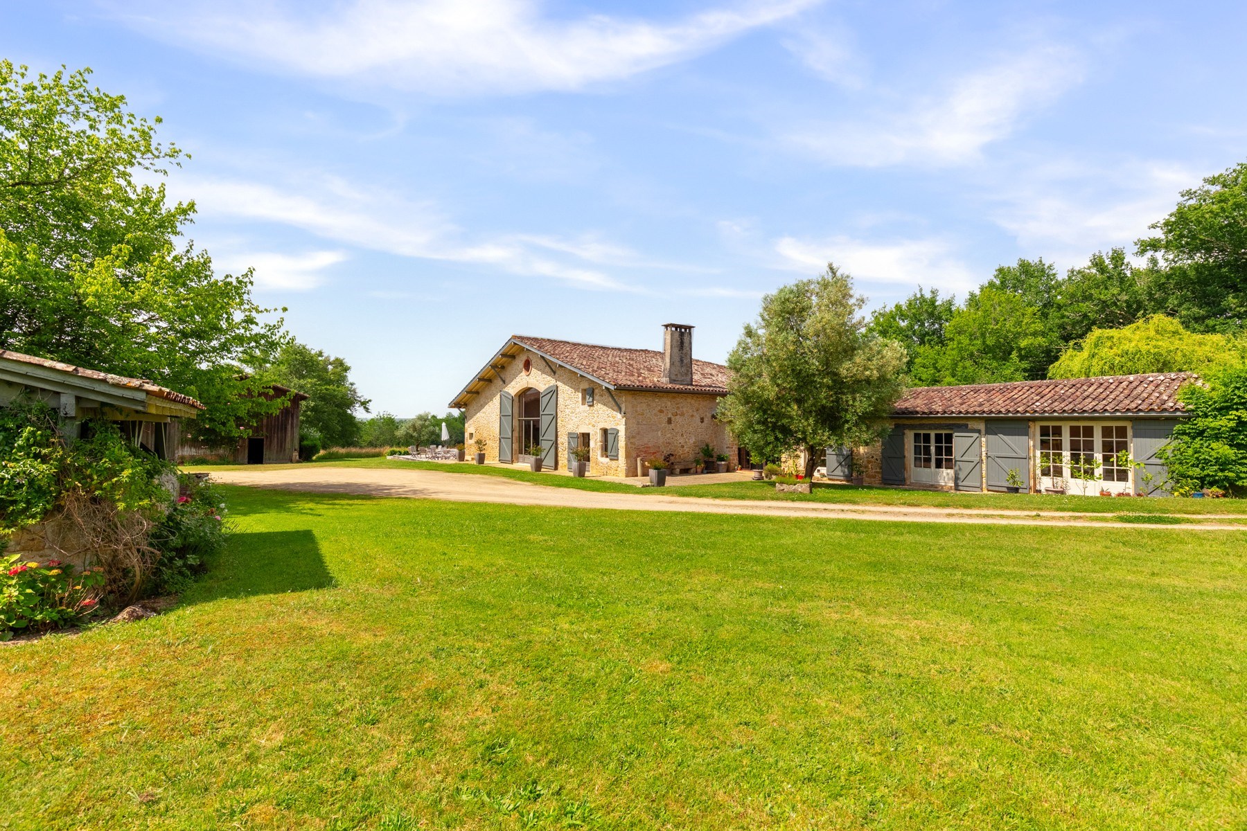 Stone country house with a red tile roof, flanked by outbuildings across a broad green lawn.