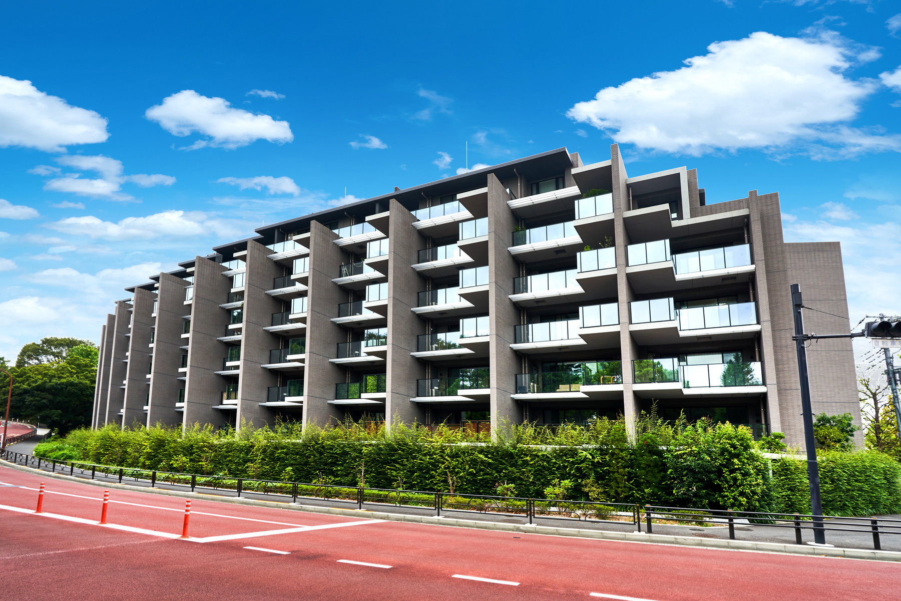 Modern apartment building with projecting balconies and landscaped hedges beside a red-paved street.