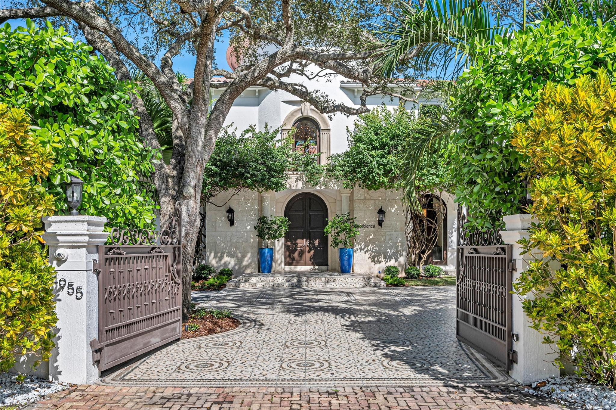 Luxury home with open wrought-iron gates, a patterned tiled driveway, and a tree-shaded arched entrance.