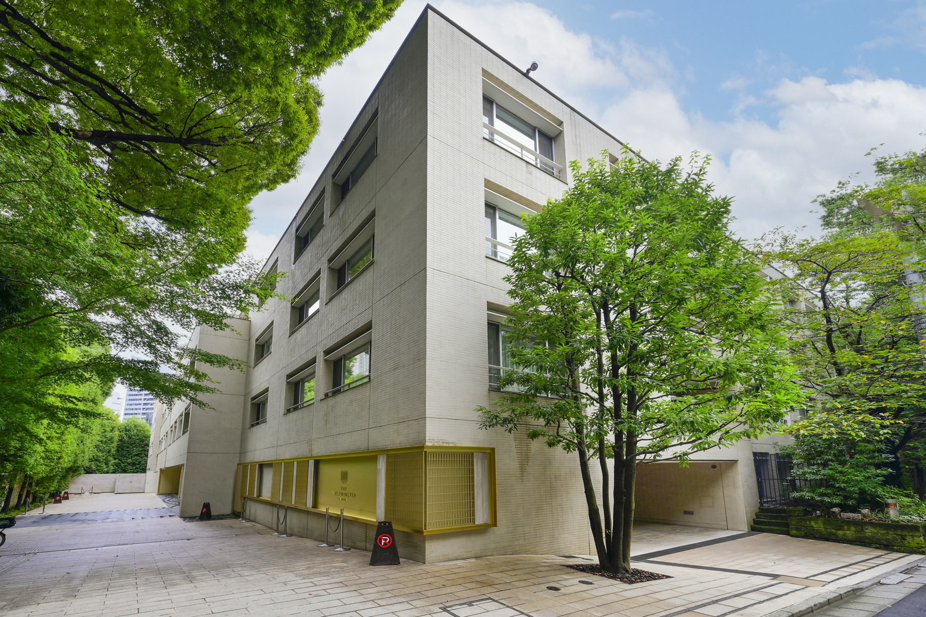 Modern tiled residential building framed by leafy trees along a paved courtyard.